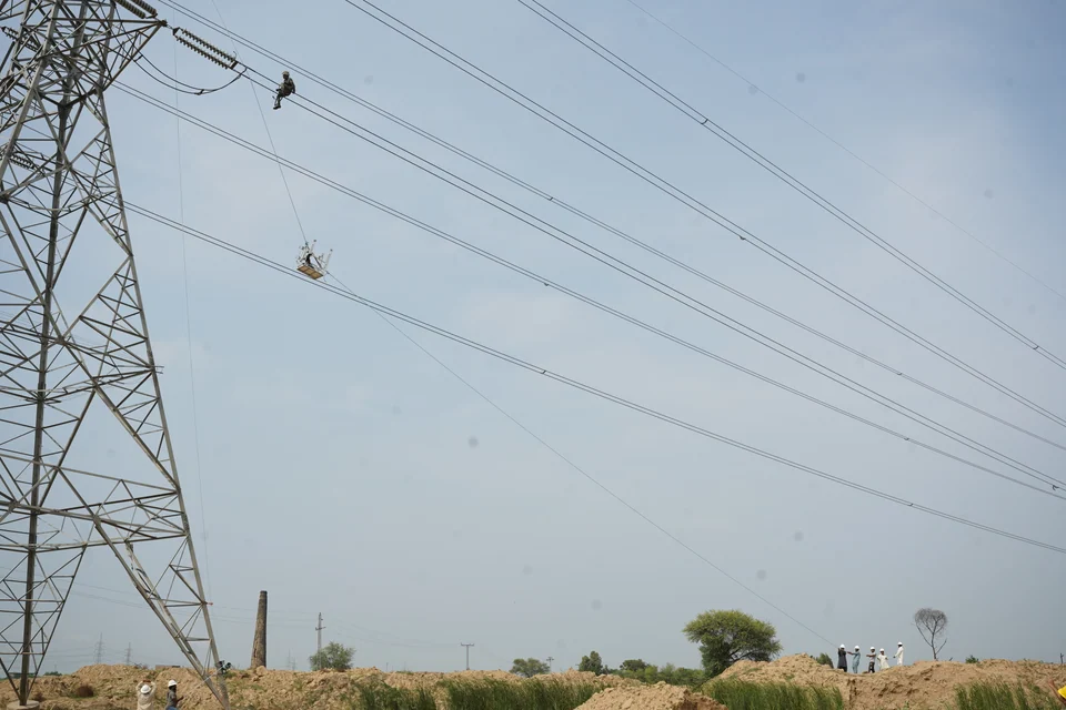 Linemen working at height on transmission conductors beside a steel tower, with a suspended work basket and colleagues on the ground