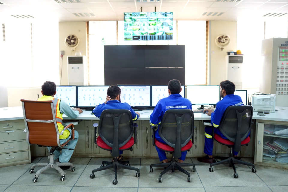 Four people at control-room workstations, three in blue workwear